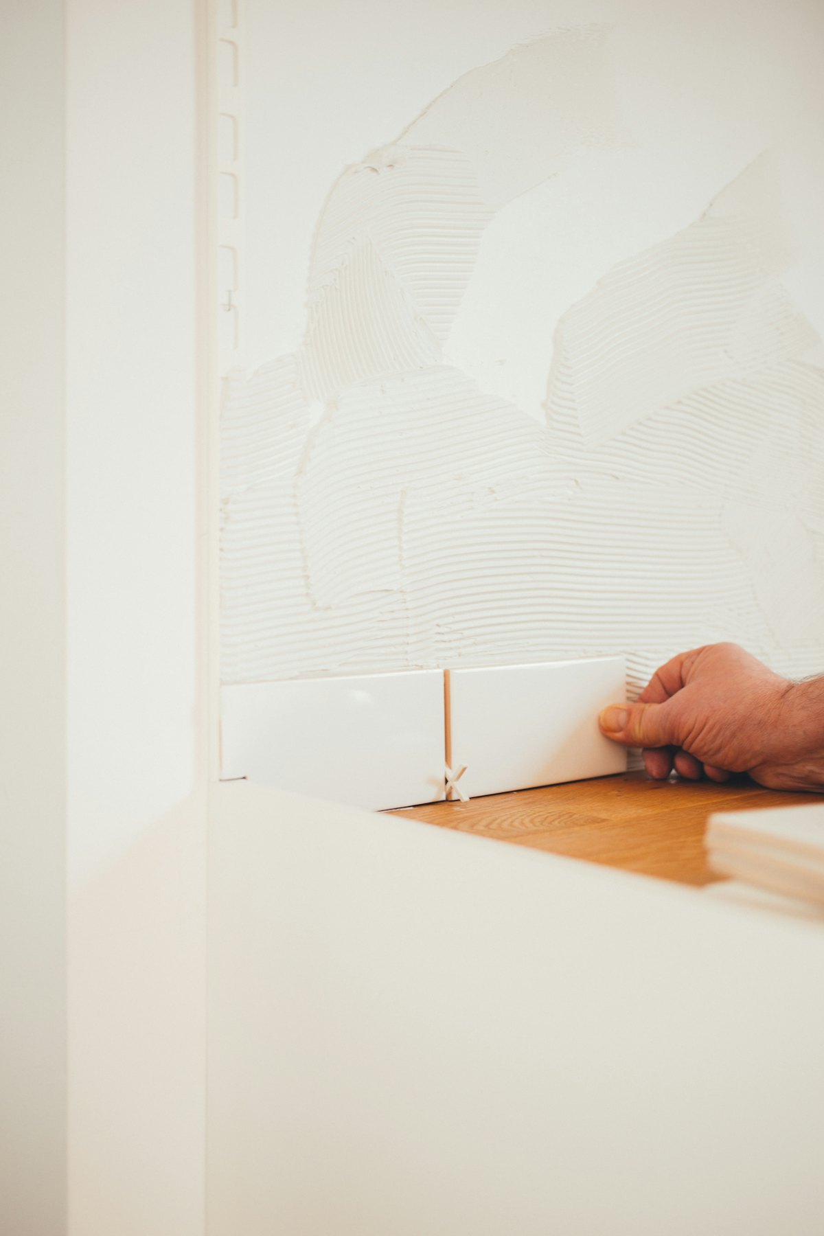 Een hand drukt een witte tegel aan op een wand met tegellijm